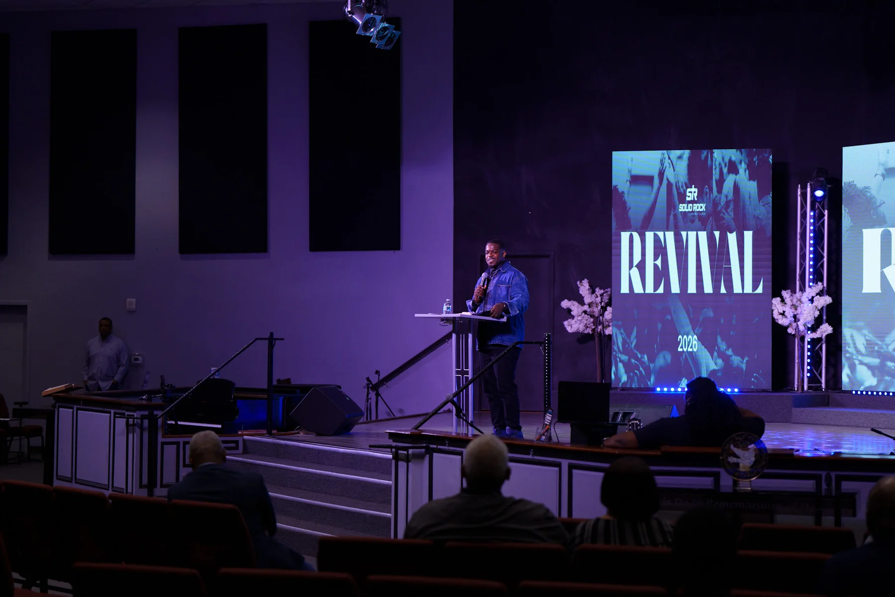 A preacher in a denim jacket speaks from the pulpit under purple stage lighting, with a Revival 2026 screen behind him and the congregation seated in front