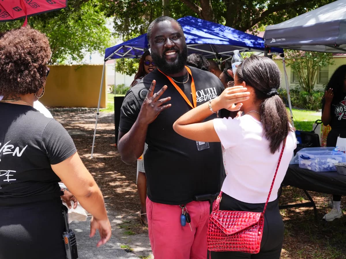 A man in a Youth Revival shirt and a staff lanyard talking and laughing with two women under event tents at an outdoor Solid Rock gathering