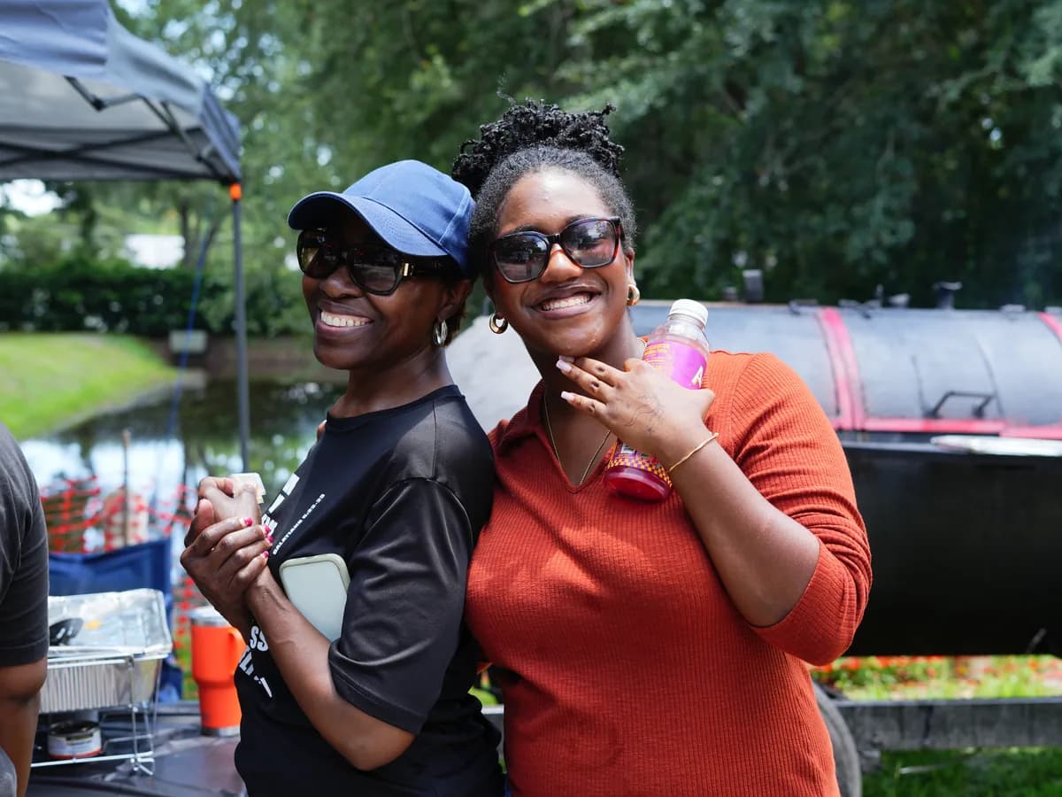 Two women with their arms around each other outdoors beside a barbecue smoker and trays of food at a Solid Rock cookout