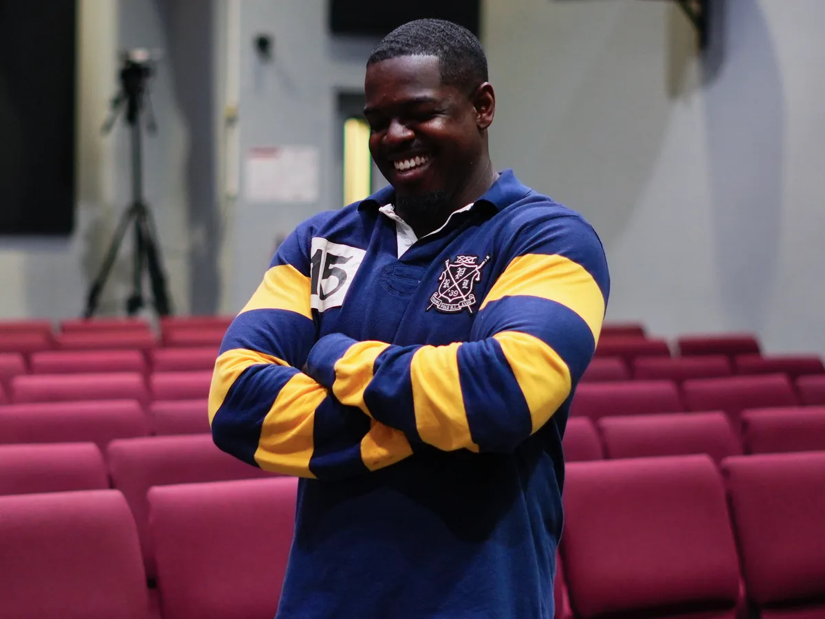 A man in a navy and gold rugby shirt laughing with his arms folded, standing in front of rows of empty maroon auditorium seats with a video camera on a tripod behind him