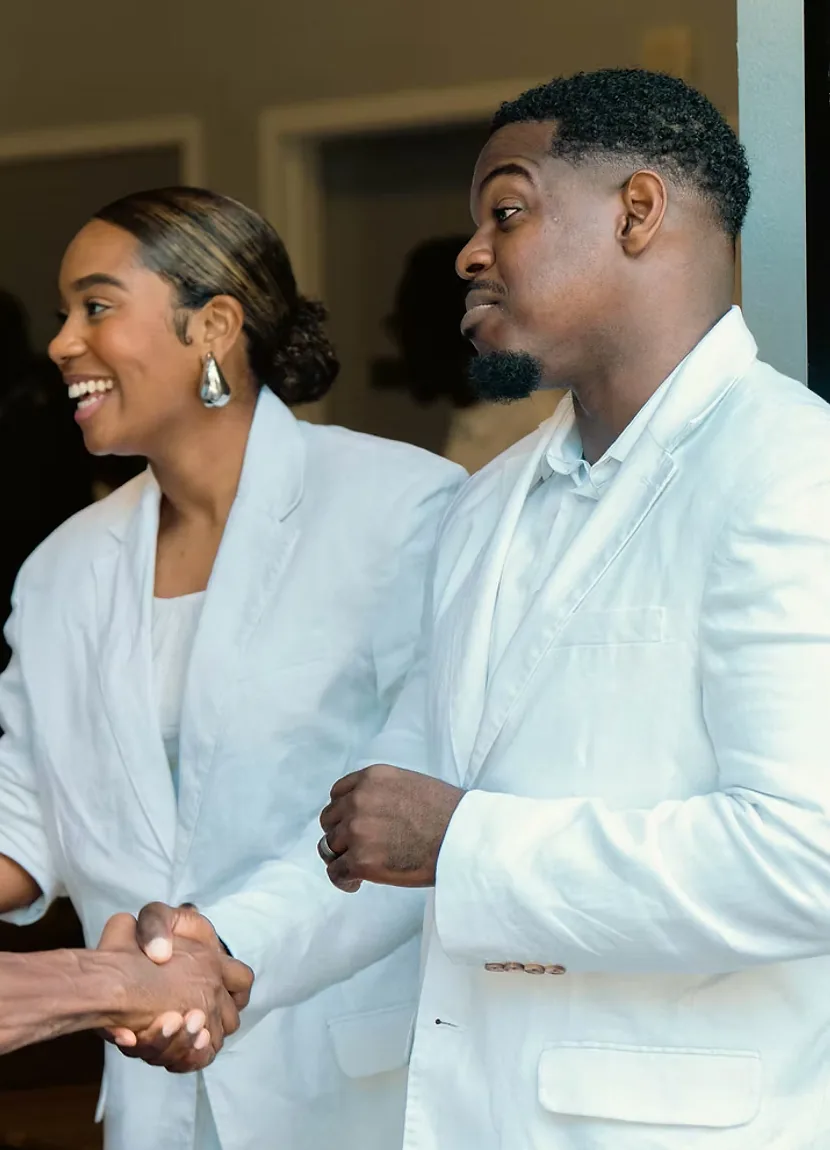 Pastor Tim and Pastor Jill Quainoo, both dressed in white, smiling as they greet a member with a handshake